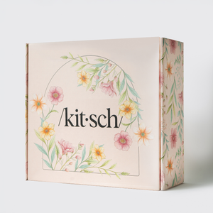 KITSCH Mother's Day Box gift box close-up — front and side view of blush pink /kit·sch/ gift box with watercolor floral wreath design featuring pink, orange, and yellow flowers with green botanical leaves — premium Mother's Day hair & beauty gift set packaging