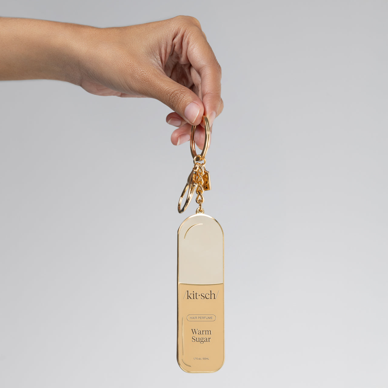Hand holding a metal enamel keychain depicting the kitsch Warm Sugar hair perfume with gold outline, design, and hardware against a gray background