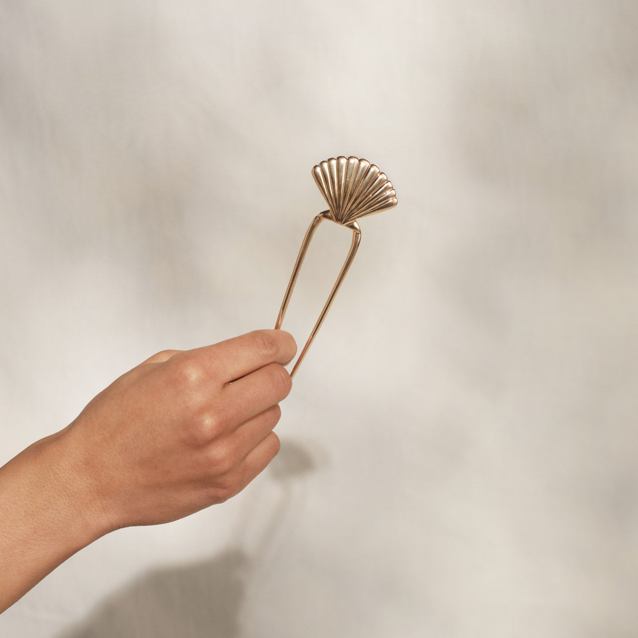 Hand holding KITSCH Metal Clam Shell French Hair Pin Gold by its prong — close-up product detail shot against warm cream background — gold U-shaped pin with fan clam shell top, full pin visible — Secure Hold | 1PC — comfortably holds hair in place without damage
