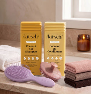 Kitsch Moisturizing Coconut Shampoo and Conditioner bottles on a bathroom counter, with a free soft violet wet/dry brush, a terracotta scalp exfoliator, and the rosewood net sponge as a free welcome kit.